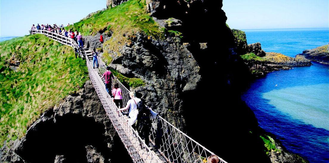 Visitors make their way across a rope bridge spanning a gap in a rocky shore area in Ireland